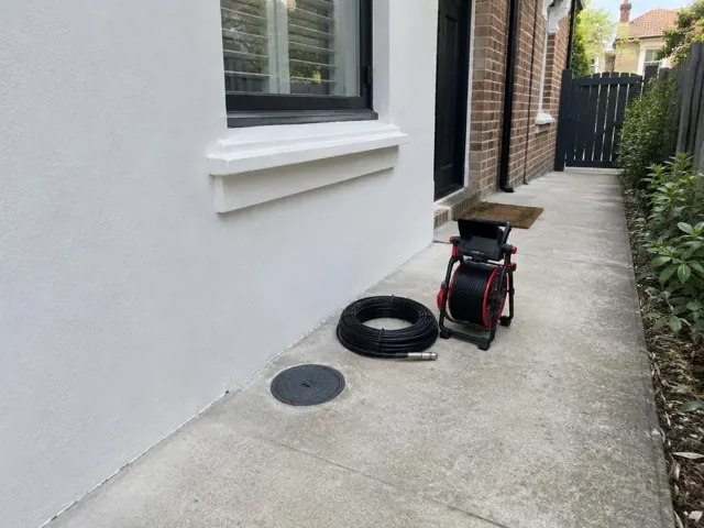 Capped drain inspection opening beside a coiled camera reel on a concrete side path by a brick house in Newtown