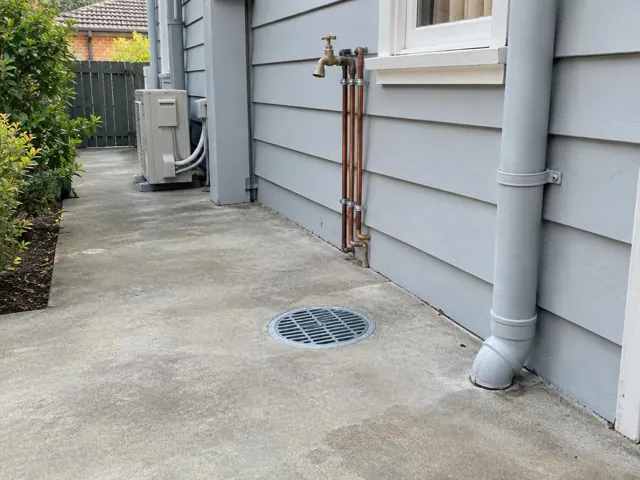 Outdoor drain grate beside grey weatherboard wall and concrete paving in Stanmore