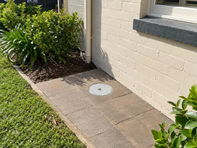 Capped access point set flush in paved side path beside a cream brick house and garden bed in Annandale